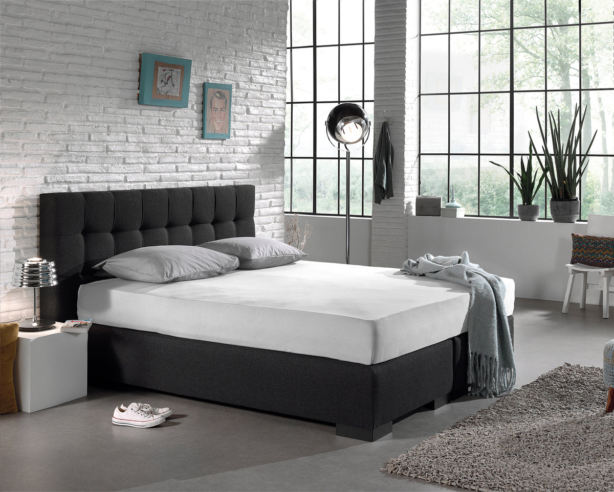 Wit hoeslaken strak om een boxspring met donkergrijs hoofdbord in een industriële slaapkamer met baksteenwand