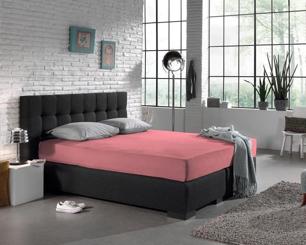 Roze jersey hoeslaken op een antracietkleurige boxspring met grijze kussens in een slaapkamer met witte bakstenen muur