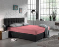 Roze jersey hoeslaken op een antracietkleurige boxspring met grijze kussens in een slaapkamer met witte bakstenen muur