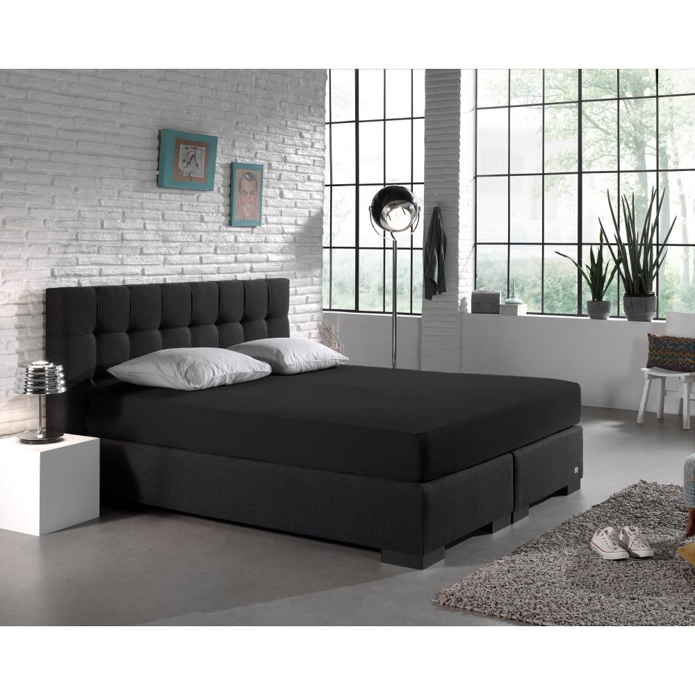 Zwart hoeslaken op een boxspring met donker gecapitonneerd hoofdbord en grijze kussens in een industriële slaapkamer