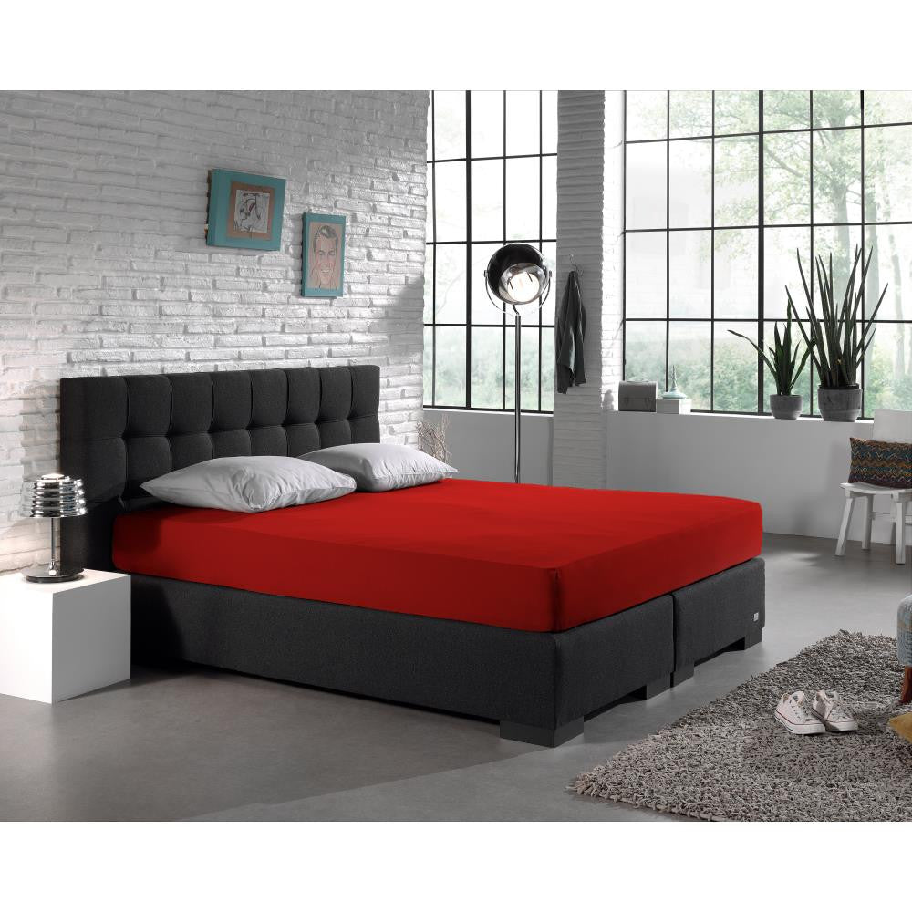 Rood hoeslaken op een boxspring met donker hoofdbord en twee grijze kussens in een slaapkamer met bakstenen muur