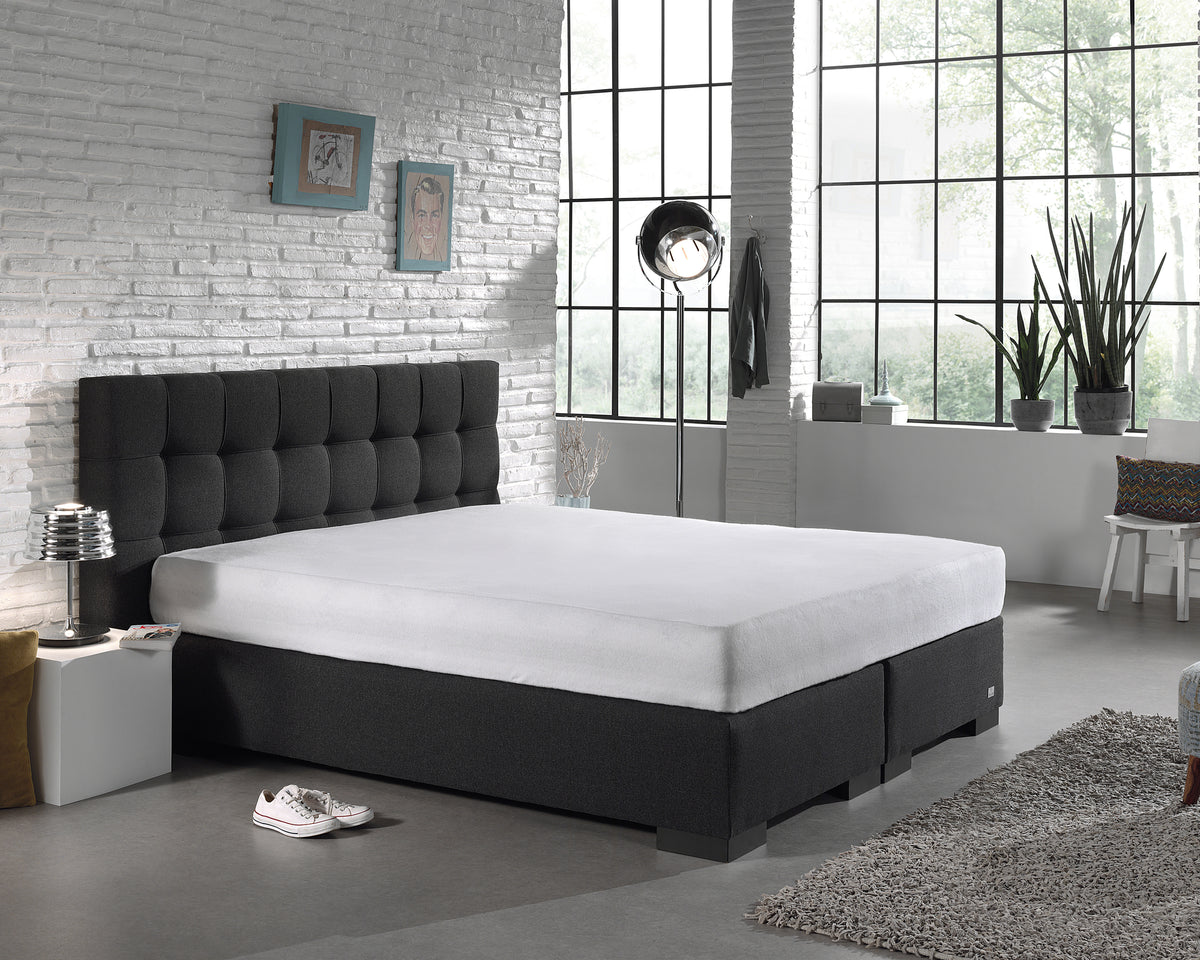 Wit molton hoeslaken op een matras met donkergrijs boxspringbed en gecapitonneerd hoofdbord in een slaapkamer
