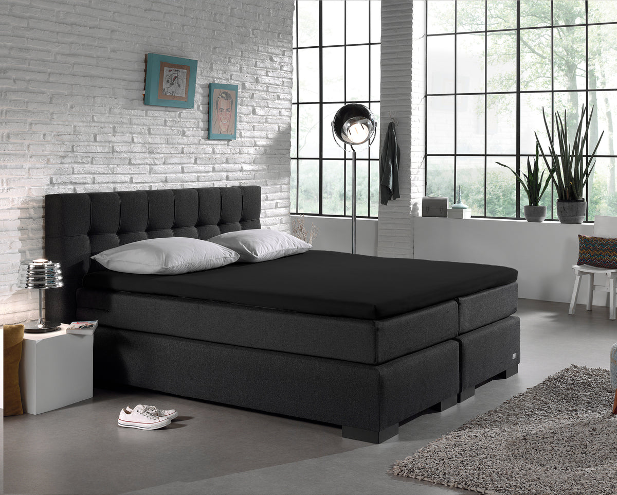 Zwart jersey hoeslaken op een donkere boxspring met grijze kussens in een lichte slaapkamer met witte baksteenmuur