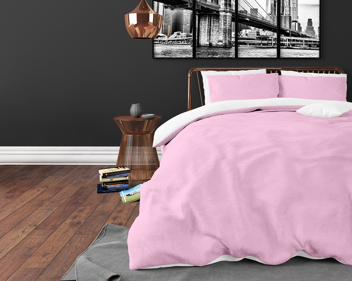 Bed met roze dekbedovertrek en roze kussenslopen met wit binnenwerk, in een donkere slaapkamer met houten vloer