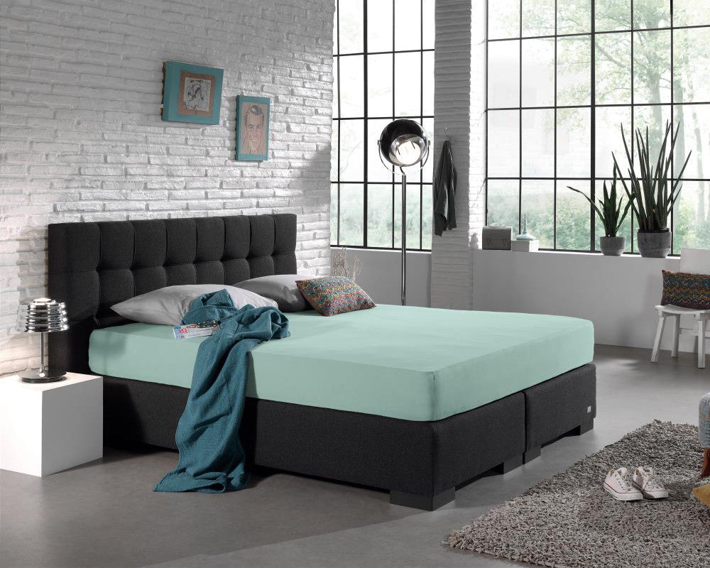 Pastelblauw hoeslaken op een boxspring met donkergrijs gecapitonneerd hoofdbord in een industriële slaapkamer