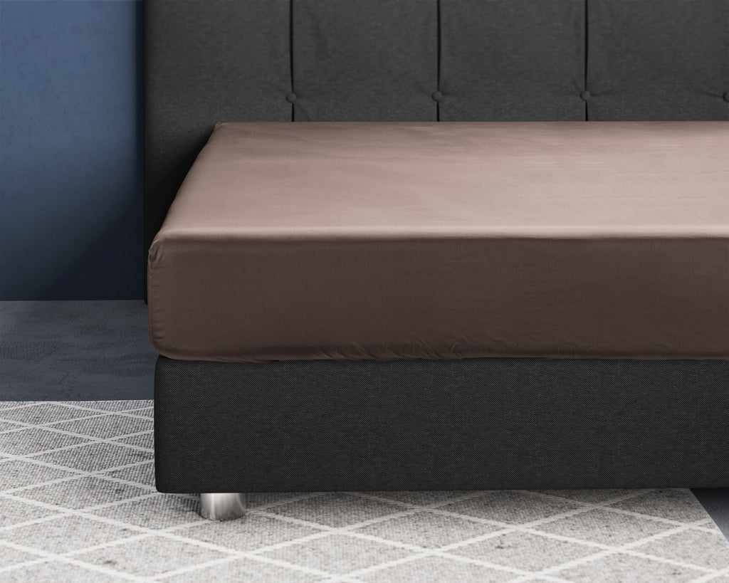 Taupe hoeslaken van satijn strak om een matras op een donkergrijs boxspringbed met gestoffeerd hoofdbord