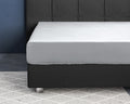 Wit satijnen hoeslaken strak om een matras op een donkergrijs bed met gecapitonneerd hoofdbord