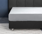 Wit satijnen hoeslaken strak om een matras op een donkergrijs bed met gecapitonneerd hoofdbord