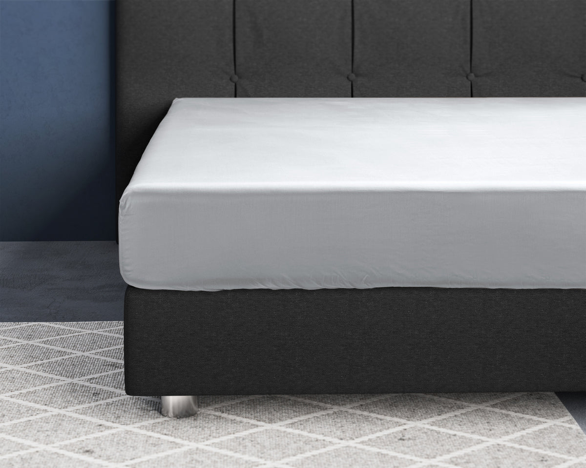 Wit satijnen hoeslaken strak om een matras op een donkergrijs bed met gecapitonneerd hoofdbord