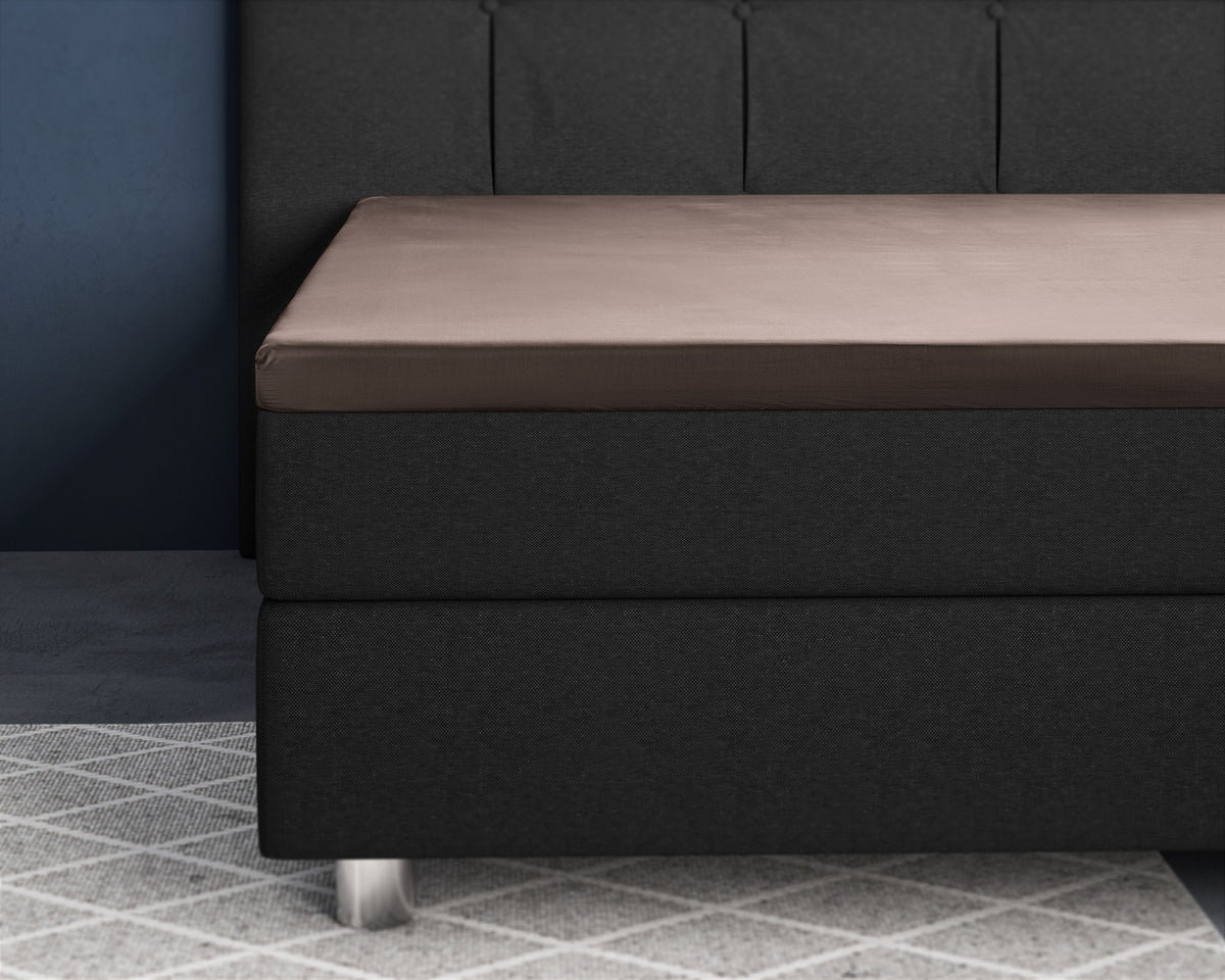 Taupe satijnen hoeslaken over een topper op een donkergrijs boxspringbed met gecapitonneerd hoofdbord
