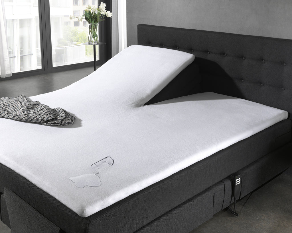 Witte waterdichte matrasbeschermer op een verstelbaar bed met donkergrijs hoofdbord, met omgevallen glas en gemorst water