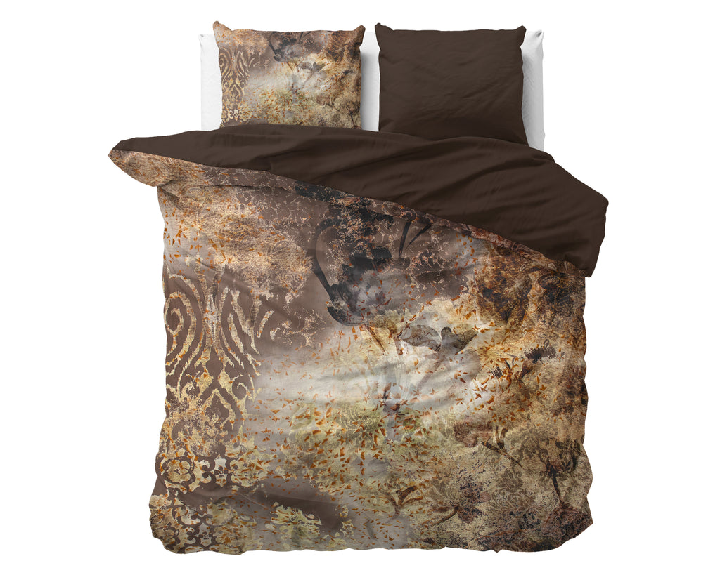 Dekbedovertrek met taupe print en goudkleurige accenten, omgeslagen met effen bruine binnenzijde en twee kussens op bed