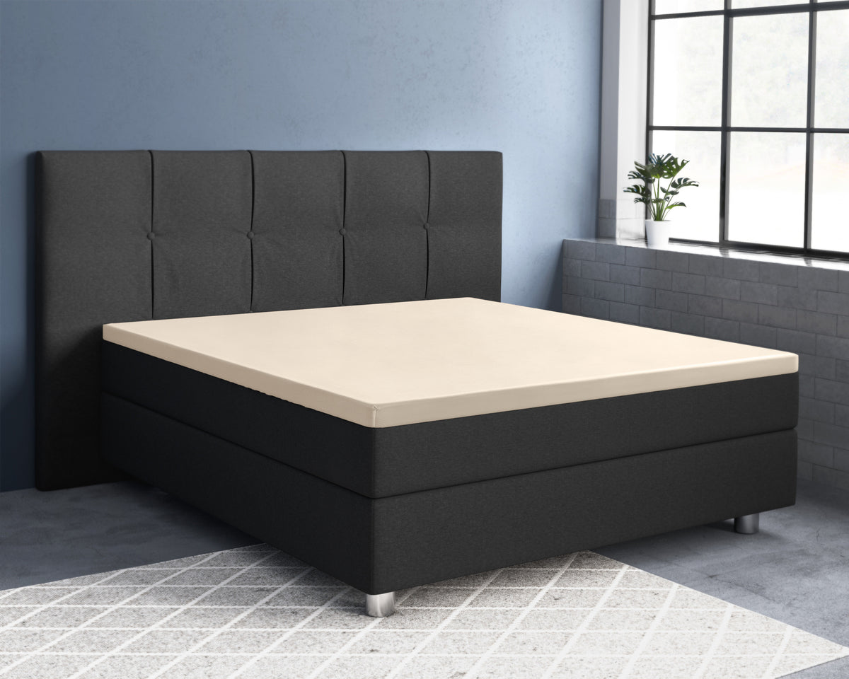 Zandkleurig hoeslaken over een topper op een donkergrijs boxspringbed met gestoffeerd hoofdbord