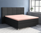 Roze satijnen hoeslaken op een oplegmatras van een zwart bed met gestoffeerd hoofdbord in een blauwgrijze slaapkamer