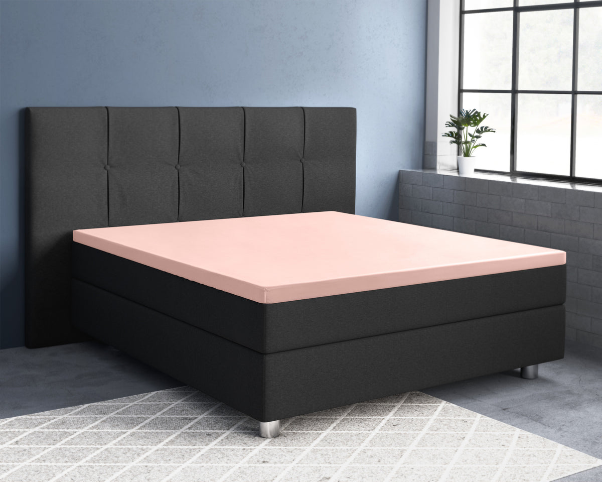 Roze satijnen hoeslaken op een oplegmatras van een zwart bed met gestoffeerd hoofdbord in een blauwgrijze slaapkamer