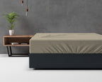 Taupe hoeslaken op een bed met donkere bekleding, naast een houten nachtkastje en een plant tegen een betonnen wand