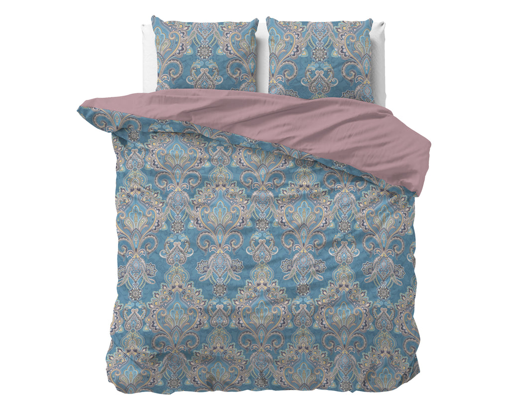 Blauw dekbedovertrek met ornamentprint en oudroze achterzijde, met twee bijpassende kussens op een bed