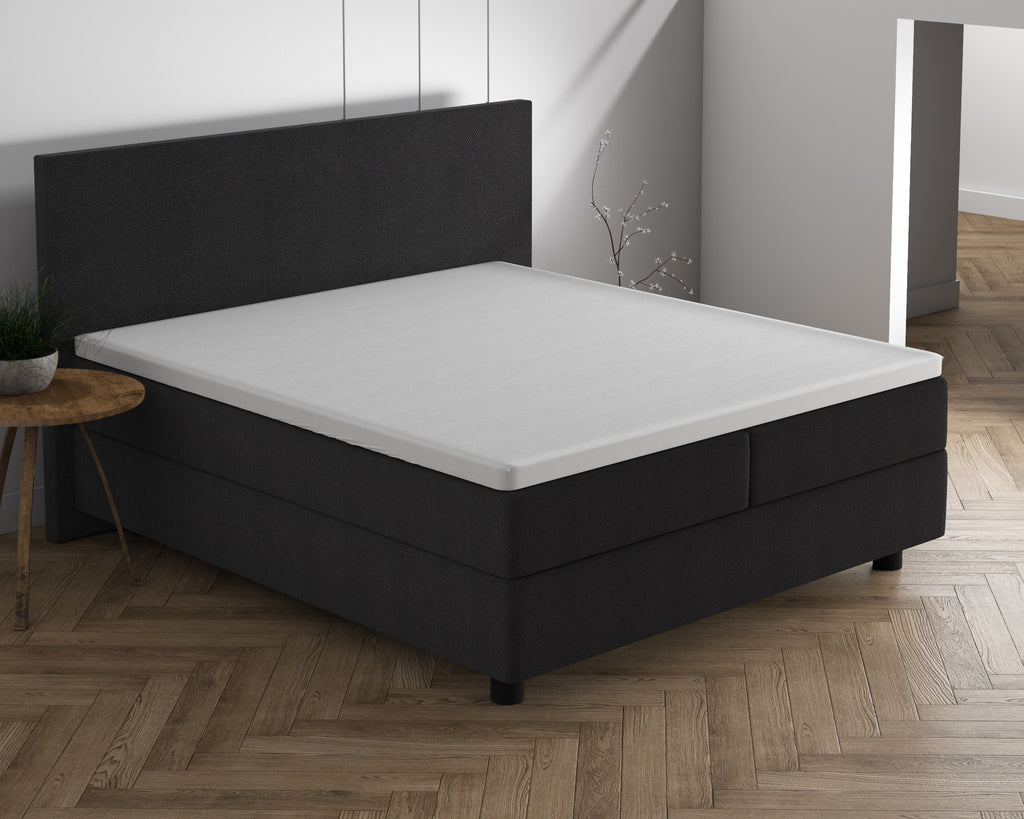 Wit topperhoeslaken op een matras van een donkergrijs boxspringbed met hoofdbord in een lichte slaapkamer