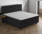 Wit topperhoeslaken op een matras van een donkergrijs boxspringbed met hoofdbord in een lichte slaapkamer
