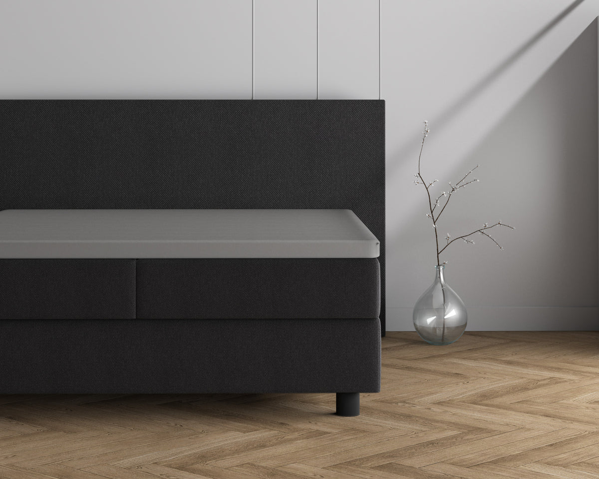 Grijs topper hoeslaken op een donkergrijs boxspringbed naast een glazen vaas met takje op een visgraatvloer