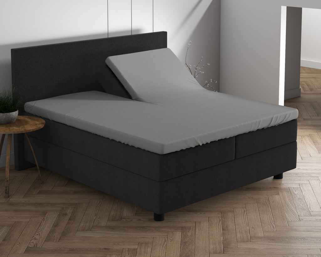 Grijs split-topper hoeslaken op een boxspring met donker hoofdbord, één helft van de topper omhoog gezet