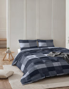Opgemaakt bed met blauw geblokt lazy dekbed en twee bijpassende kussens, met grijs plaid in een lichte slaapkamer