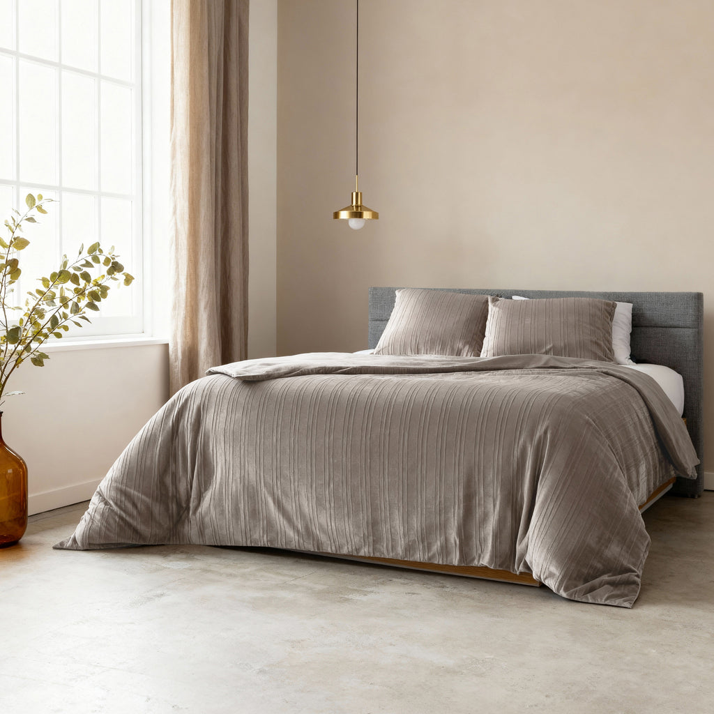 Bed opgemaakt met taupe/grijs fluwelen dekbedovertrek en kussens, onder een gouden hanglamp in een lichte slaapkamer