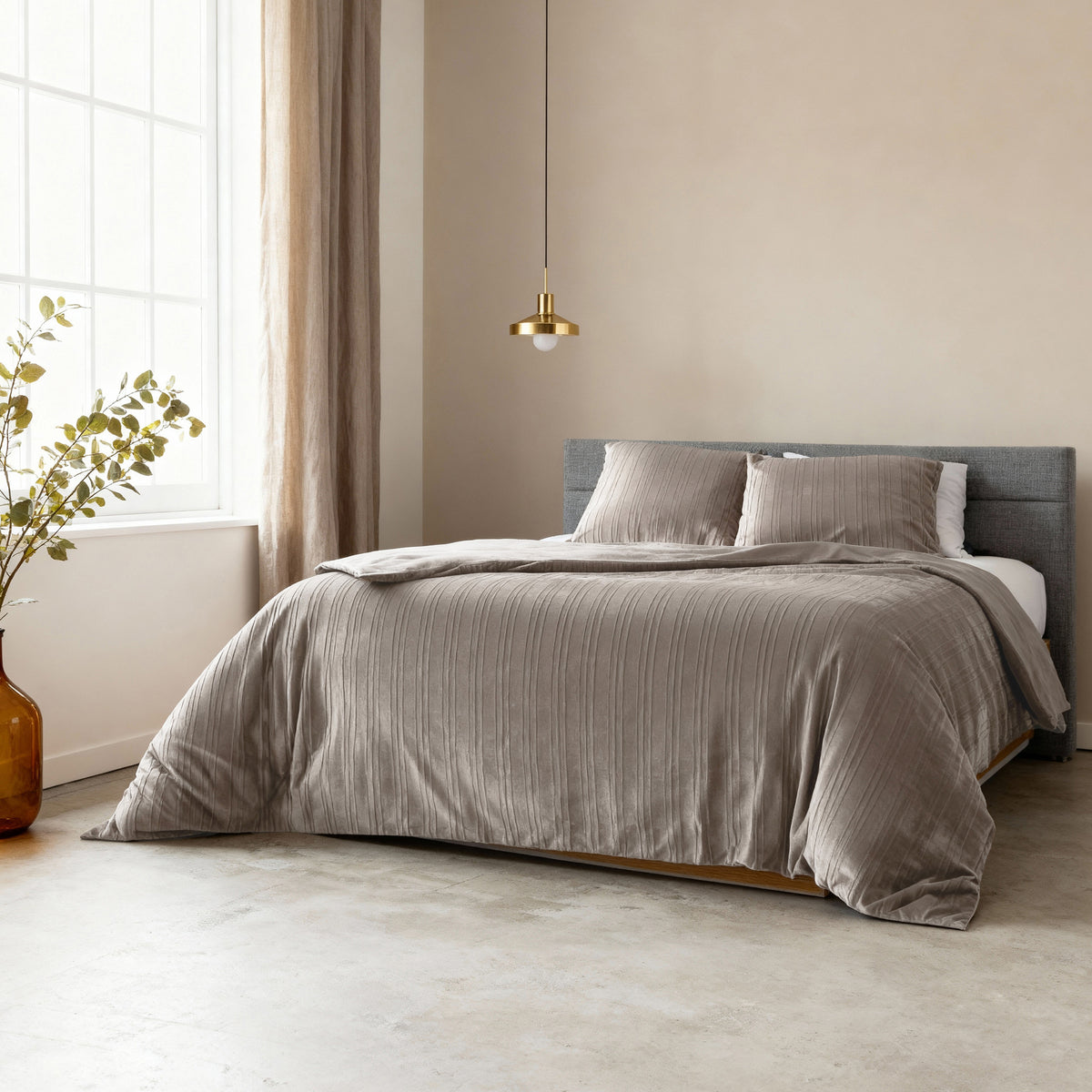 Bed opgemaakt met taupe/grijs fluwelen dekbedovertrek en kussens, onder een gouden hanglamp in een lichte slaapkamer