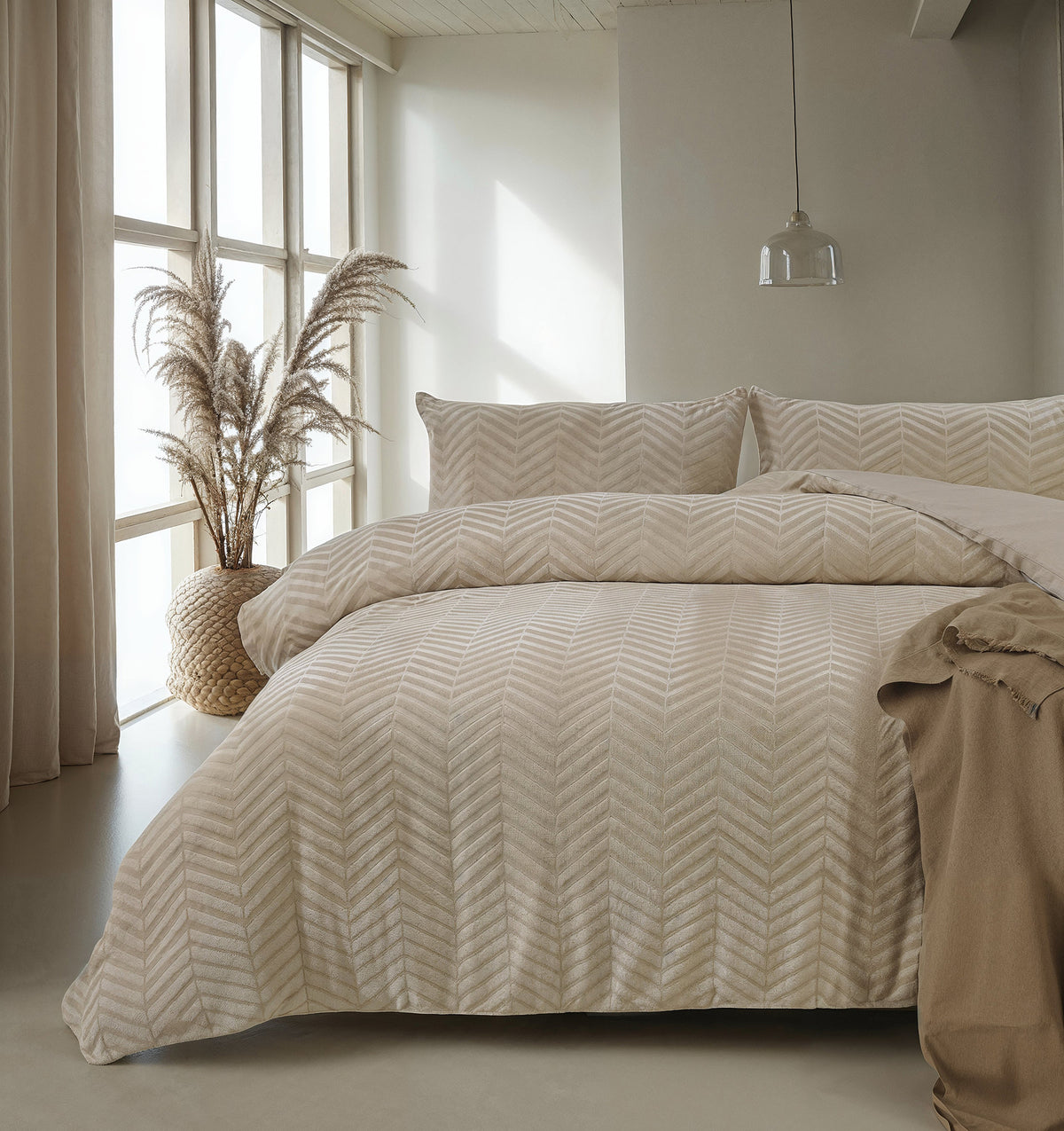 Bed met taupe teddy dekbedovertrek met visgraatpatroon in een lichte slaapkamer met pampasgras en hanglamp