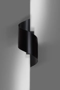 Zwarte spiraalvormige wandlamp aan een grijze muur met zacht licht dat langs de wand valt