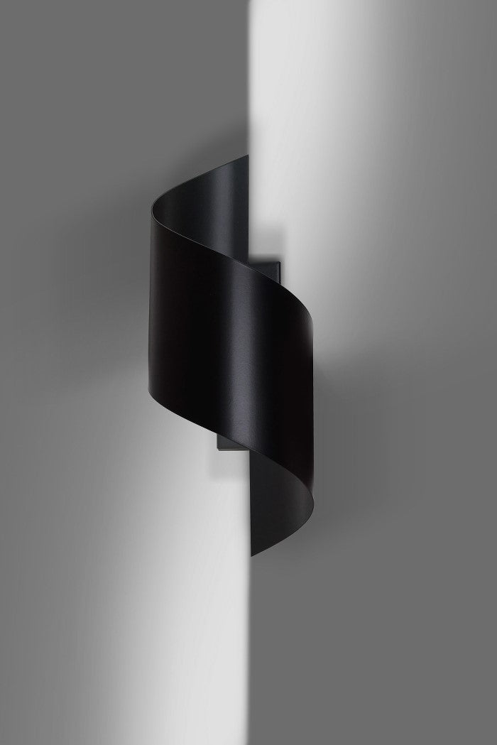 Zwarte spiraalvormige wandlamp aan een grijze muur met zacht licht dat langs de wand valt