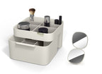 Grijze make-up organiser met vakken en lade, gevuld met kwast en make-up, naast een opengeklapte handspiegel