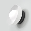 Witte wandlamp met schuin geplaatste kap, lichtgevend bolletje en zwarte ronde basis tegen een lichte wand