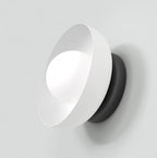 Witte wandlamp met schuin geplaatste kap, lichtgevend bolletje en zwarte ronde basis tegen een lichte wand