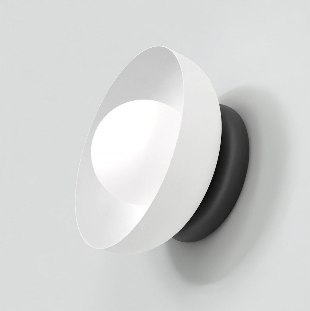 Witte wandlamp met schuin geplaatste kap, lichtgevend bolletje en zwarte ronde basis tegen een lichte wand