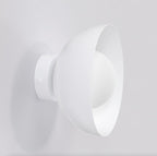 Witte wandlamp met schuin geplaatste komvormige kap en ronde wandplaat, tegen een lichte muur